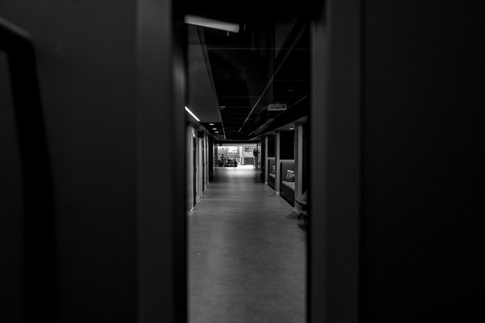 A dark corridor seen through the gap of a held-open door, daylight at the far end.