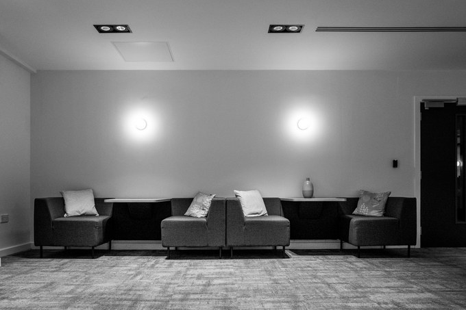 A waiting area of sofas and squared cushions under two wall lights, nobody sitting.