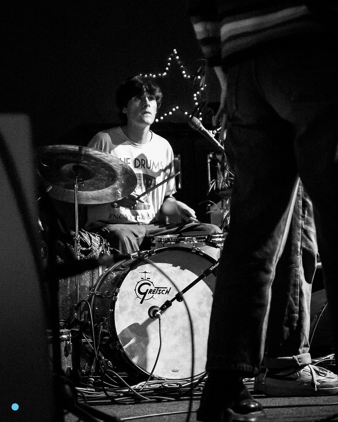 Black and white photograph of a drummer behind a Gretsch kit on a dark stage, a paper star pinned to the wall behind him, drumsticks in motion.