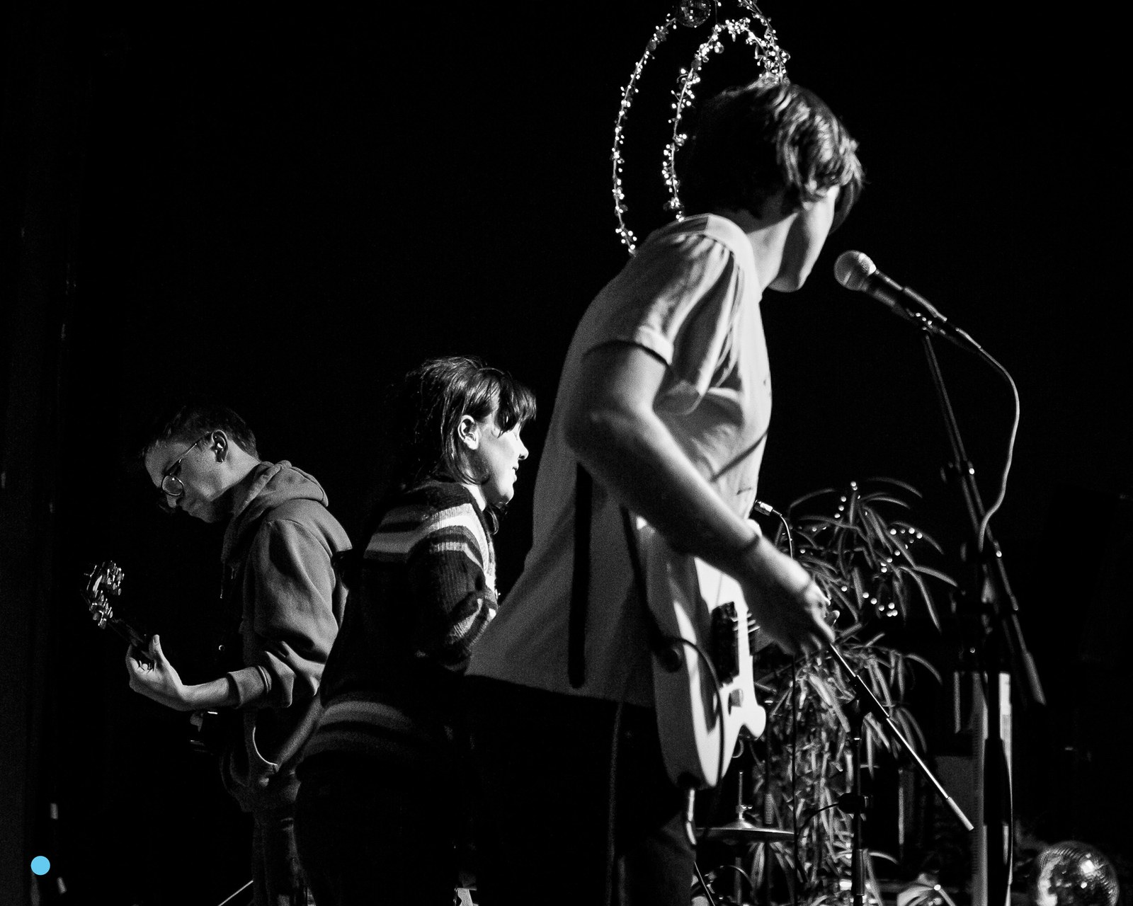Black and white photograph of the band on a small dark stage, a guitarist at the left, the singer at a central microphone with his back turned, the drummer behind, a star of fairy lights strung above.