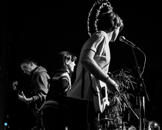 Black and white photograph of the band on a small dark stage, a guitarist at the left, the singer at a central microphone with his back turned, the drummer behind, a star of fairy lights strung above.
