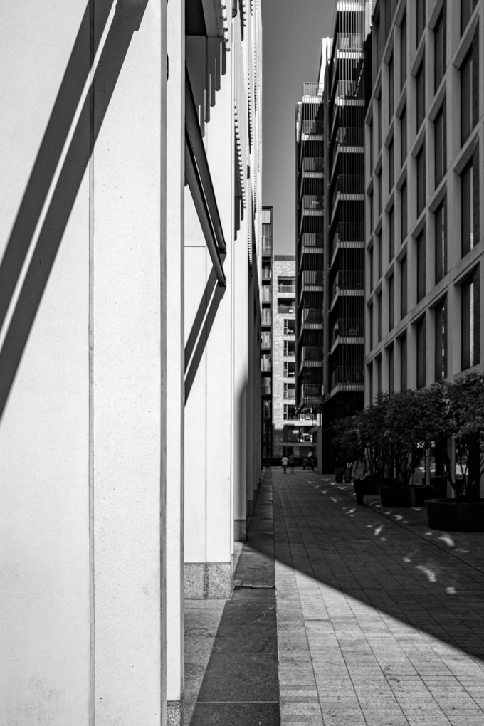 A narrow canyon between modern London buildings, a hard diagonal of shadow across pale stone, trees in planters at the far end.