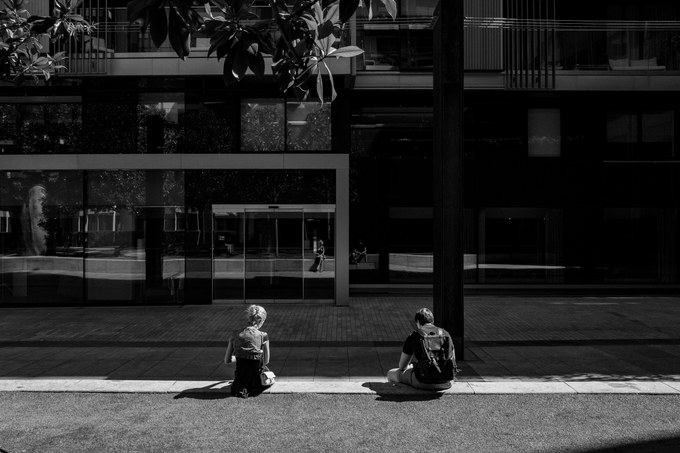 Two people sitting apart on a kerb in full sun outside a glass-fronted building, their reflections in the glass behind.