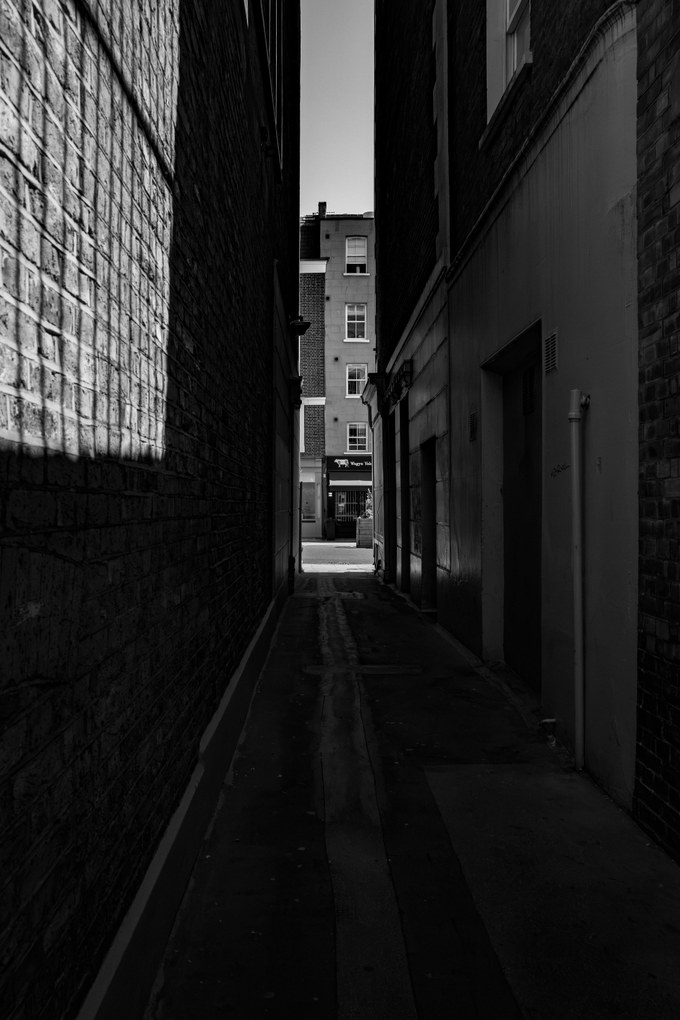 A very narrow alley between buildings, a shaft of sunlight on the brick at left, a Georgian frontage closing the far end.