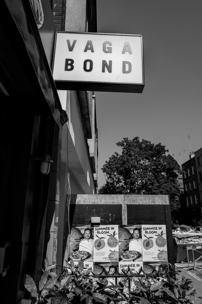 A projecting box sign reading VAGABOND above a dark shopfront, posters pasted on hoarding below and a plane tree behind.