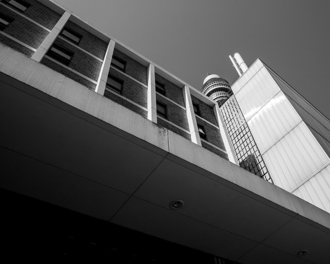 Looking up past a brick and concrete roofline to the BT Tower standing against a clear sky.