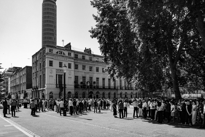 Dozens of office workers standing on the pavement and roadway at Fitzroy Square in bright sun, a Georgian terrace and the BT Tower behind them.