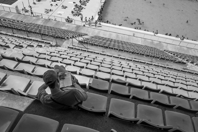 A man alone in the empty stands, cap on, reading a book, the pool and its swimmers far below him.