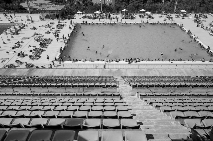 Rows of empty stand seating in the foreground, the crowded swimming pool and its sunbathers beyond the barrier.