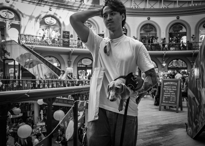 A young man in a white T-shirt holding a dachshund under one arm, hand pushed through his hair, the domed galleries of the Leeds Corn Exchange behind him.