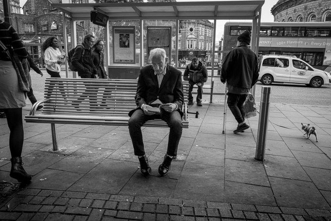 An elderly man in a suit reading on a bus shelter bench while people walk past, the Corn Exchange behind.