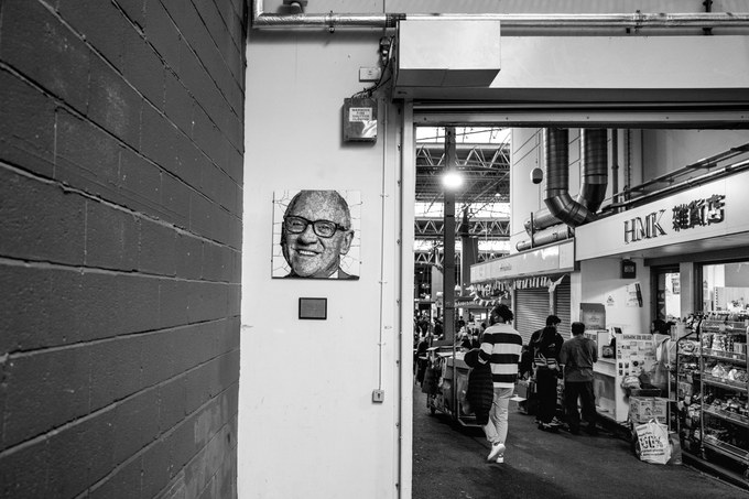 A mosaic portrait of a smiling man in glasses on a market wall, shoppers moving through the hall beyond.