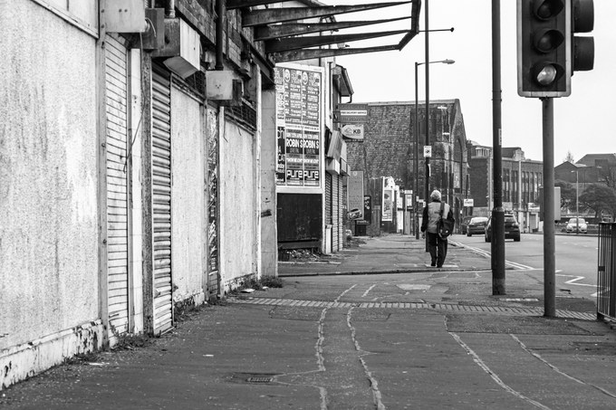 Boarded shopfronts along the New Osborne frontage on Oldham Road, a lone figure walking away.