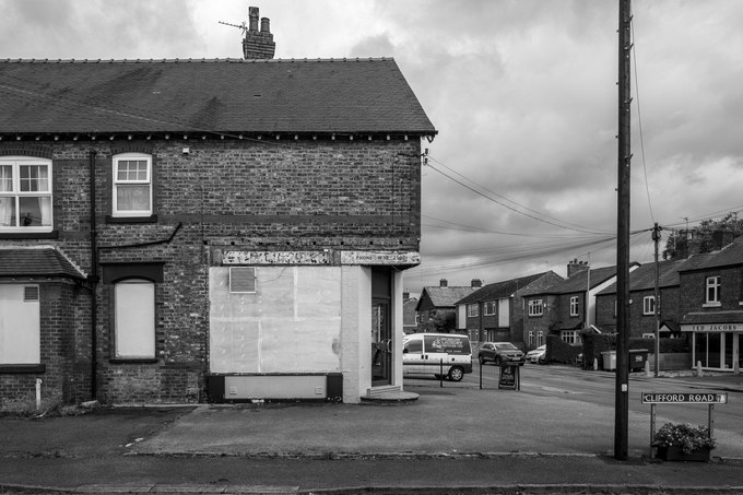 A stripped-out shop unit, the window boarded with bare panels and older ghost lettering surviving along the fascia above.